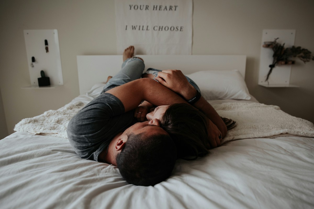 A couple lies on a bed in a cozy, intimate embrace, with a cloth banner above them that reads “YOUR HEART I WILL CHOOSE.” The room has minimal, warm-toned decor, giving a soft, romantic feel.