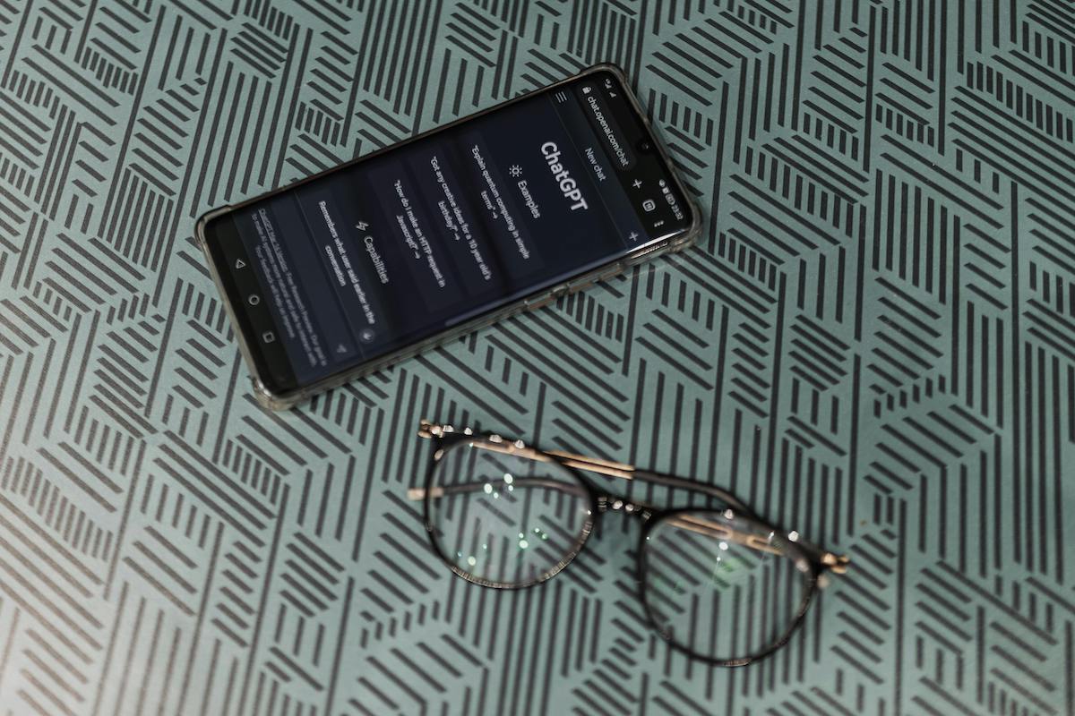 A phone with a pair of glasses. The phone displays the ChatGPT homepage. This image symbolizes the increasing use of AI tools like ChatGPT to answer questions and potentially replace traditional couples therapy.