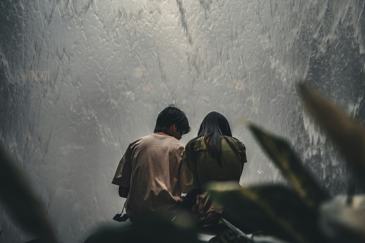 Two people sit closely together with their backs to the camera, facing a misty waterfall wall. They appear deep in conversation or quiet connection, surrounded by blurred plants in the foreground.
