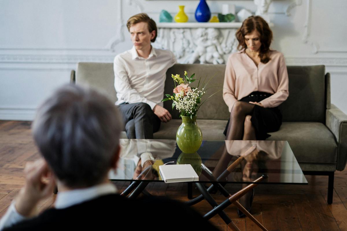 A couple sits apart on a couch during a therapy session, with a therapist in the foreground observing them. A green vase with flowers sits on a glass table between them, highlighting emotional distance.