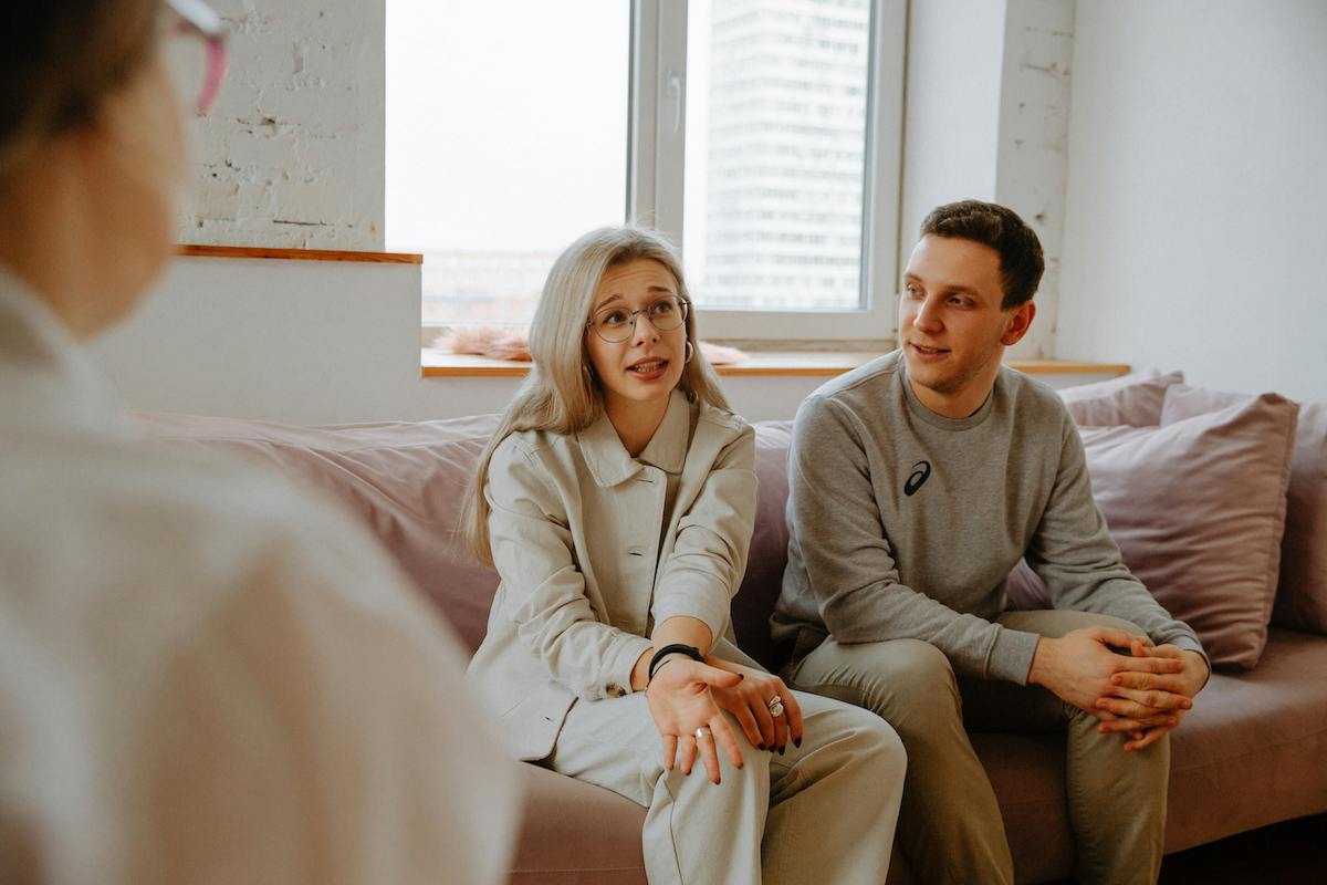 A woman with long light hair gestures while speaking during a therapy session, seated beside a man who listens attentively. The therapist, partially visible in the foreground, is engaging with them in a bright, modern room.