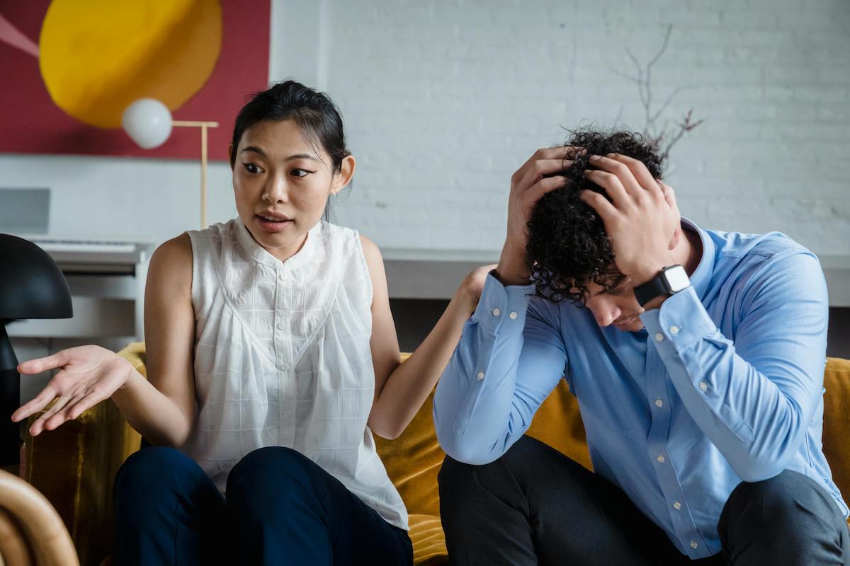 A woman looks frustrated while speaking, gesturing with one hand, as a man beside her buries his face in his hands. The modern setting and expressive body language convey relationship tension.
