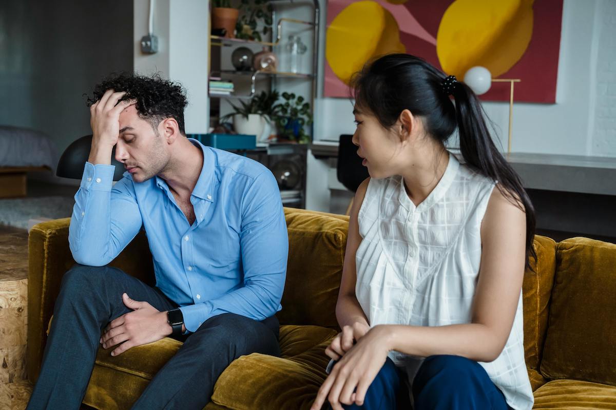 A woman sits on a yellow couch speaking with concern while a man beside her rests his head in his hand, looking stressed and overwhelmed, suggesting a relationship argument or emotional tension.