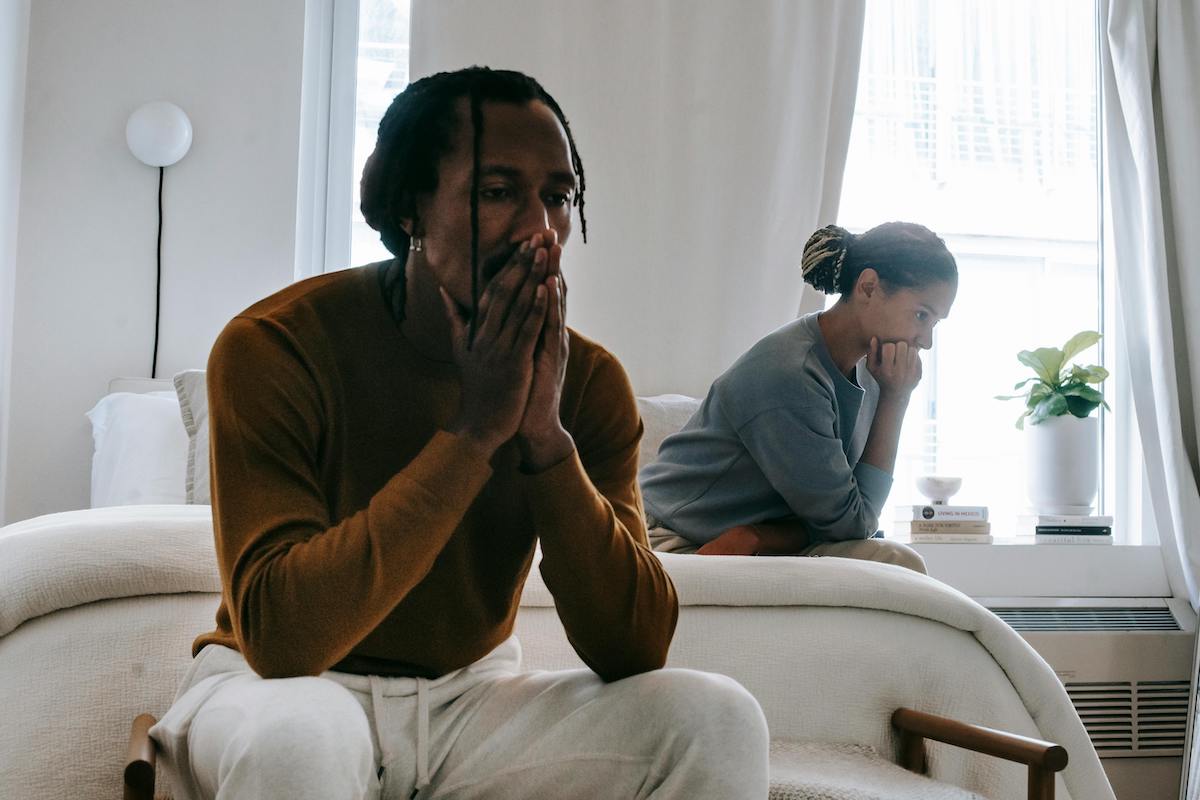 A man and woman sit in a softly lit room, both looking pensive and upset, facing away from each other with their hands near their faces in frustration. The atmosphere suggests a serious disagreement or emotional strain.