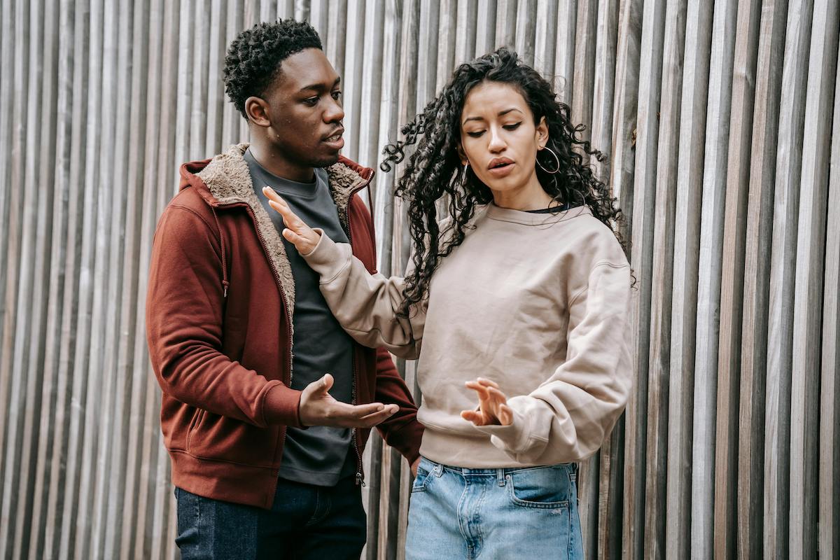 A couple argues in front of a tall wooden fence, with the woman holding up her hands in frustration while the man tries to reason with her. Their body language conveys miscommunication and tension.