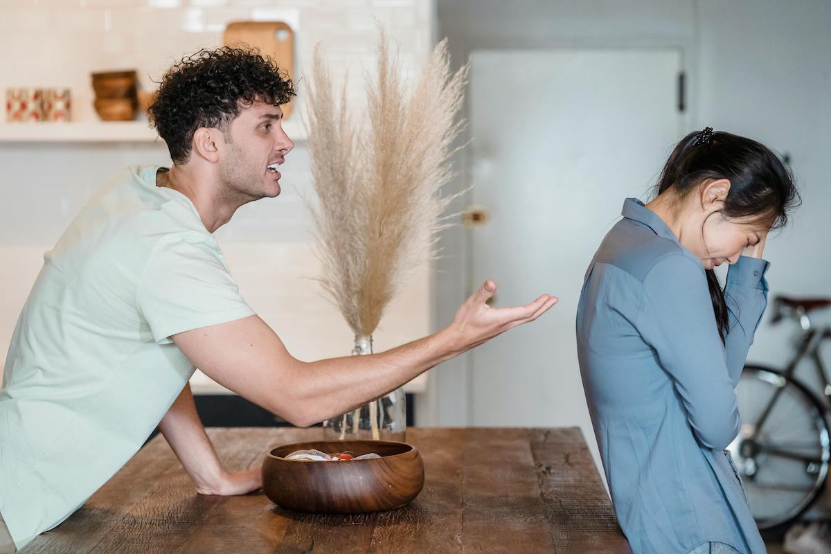 A man angrily gestures toward a woman who is turned away and visibly distressed, hiding her face in her hand. The confrontation takes place in a kitchen-like setting, emphasizing emotional conflict.