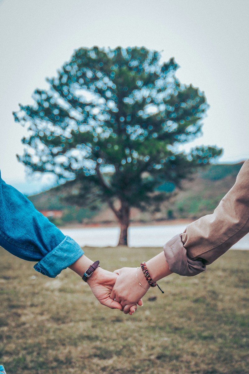 Two people hold hands outdoors with a blurred tree in the background. The focus is on the connection between their hands, symbolizing unity and support.