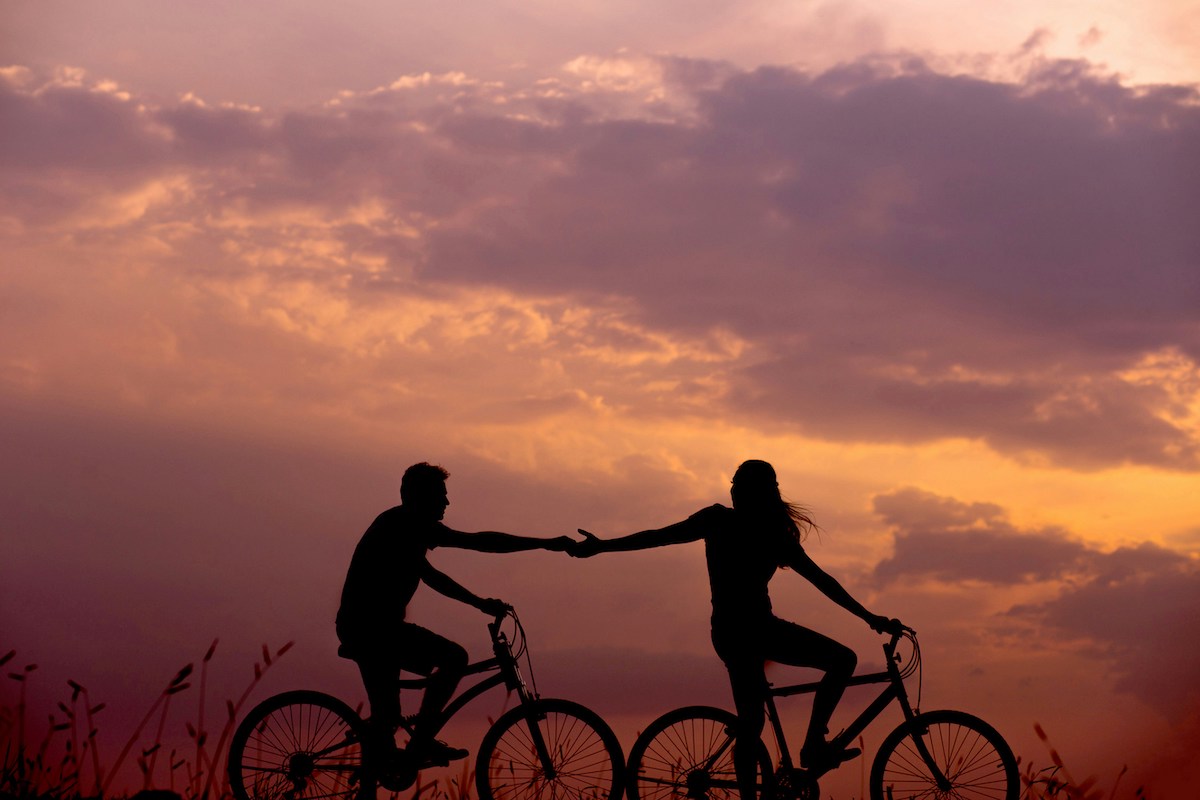 A couple rides bicycles at sunset, holding hands as they cycle side by side. The sky glows with rich purples and oranges, highlighting romance and freedom.
