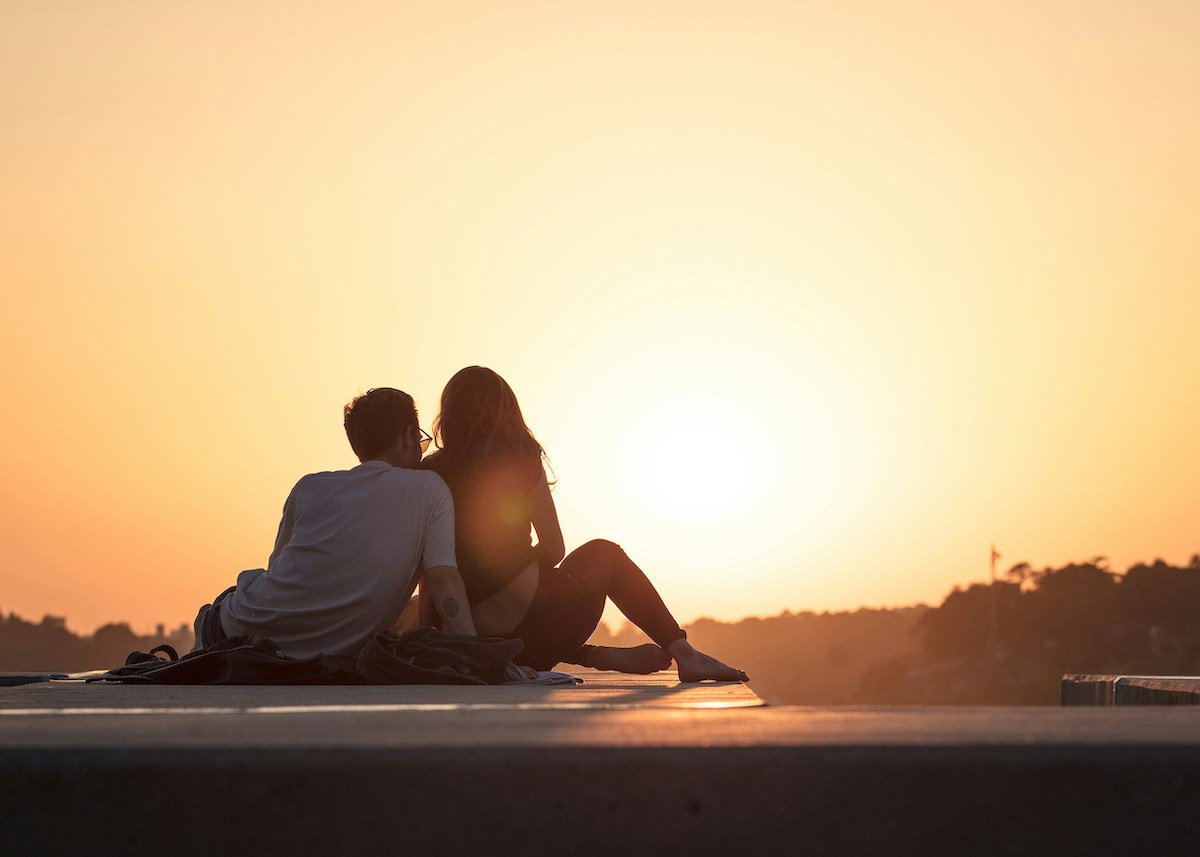 A couple sits together at sunset, gazing at the glowing horizon while seated on a ledge. Their silhouettes and close proximity evoke a sense of peace and intimacy.