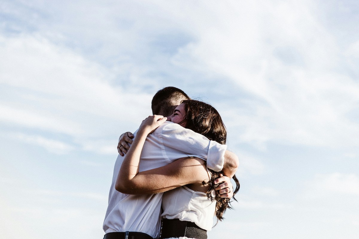 A couple embraces tightly under a bright sky, both dressed in white and smiling as they share a warm hug. The setting and their expressions radiate happiness and connection.