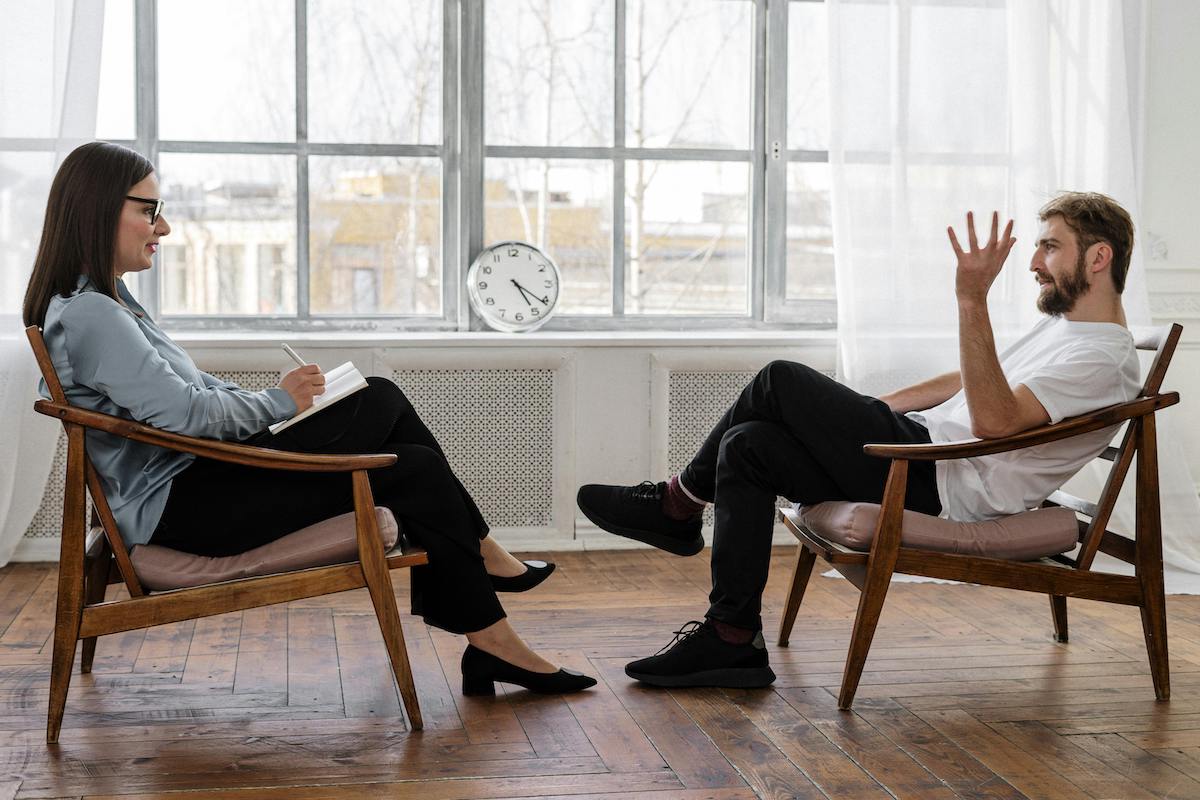 A male client gestures expressively while seated across from a female therapist who listens and takes notes in a bright, minimalistic office with wooden floors and a clock on the windowsill.
