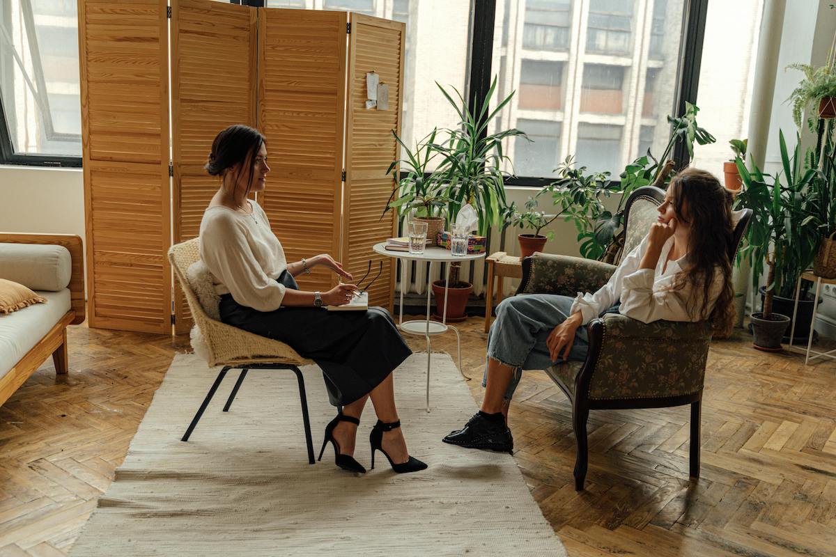 A woman sits in a therapy session, leaning forward while talking with a female therapist in a room filled with natural light and houseplants. A small table with notebooks and water glasses sits between them.