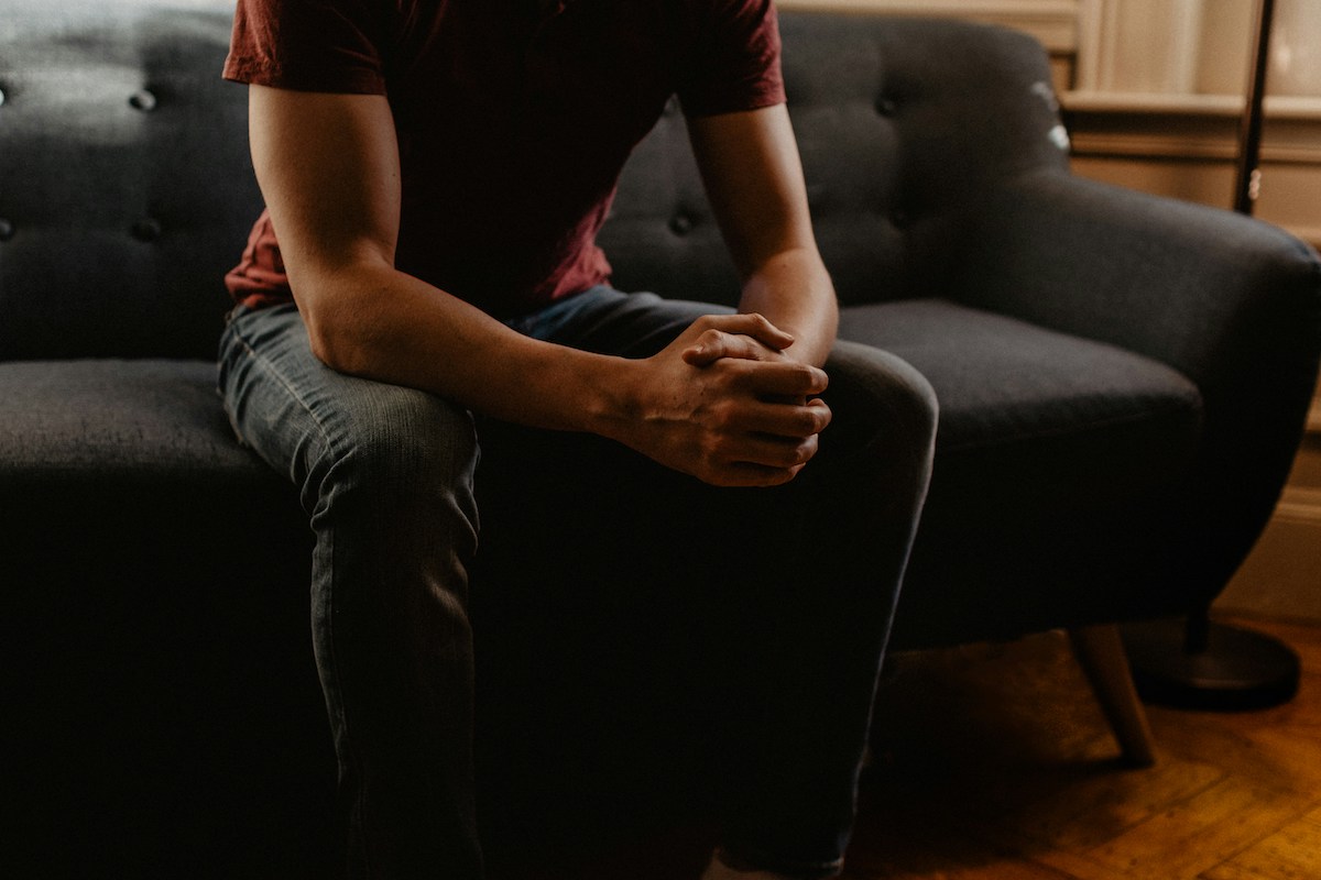 A person wearing a red shirt sits with clasped hands on a couch in a softly lit room. This image evokes the emotional support typically offered in couples therapy, raising questions about whether AI solutions like ChatGPT could provide the same level of empathy.