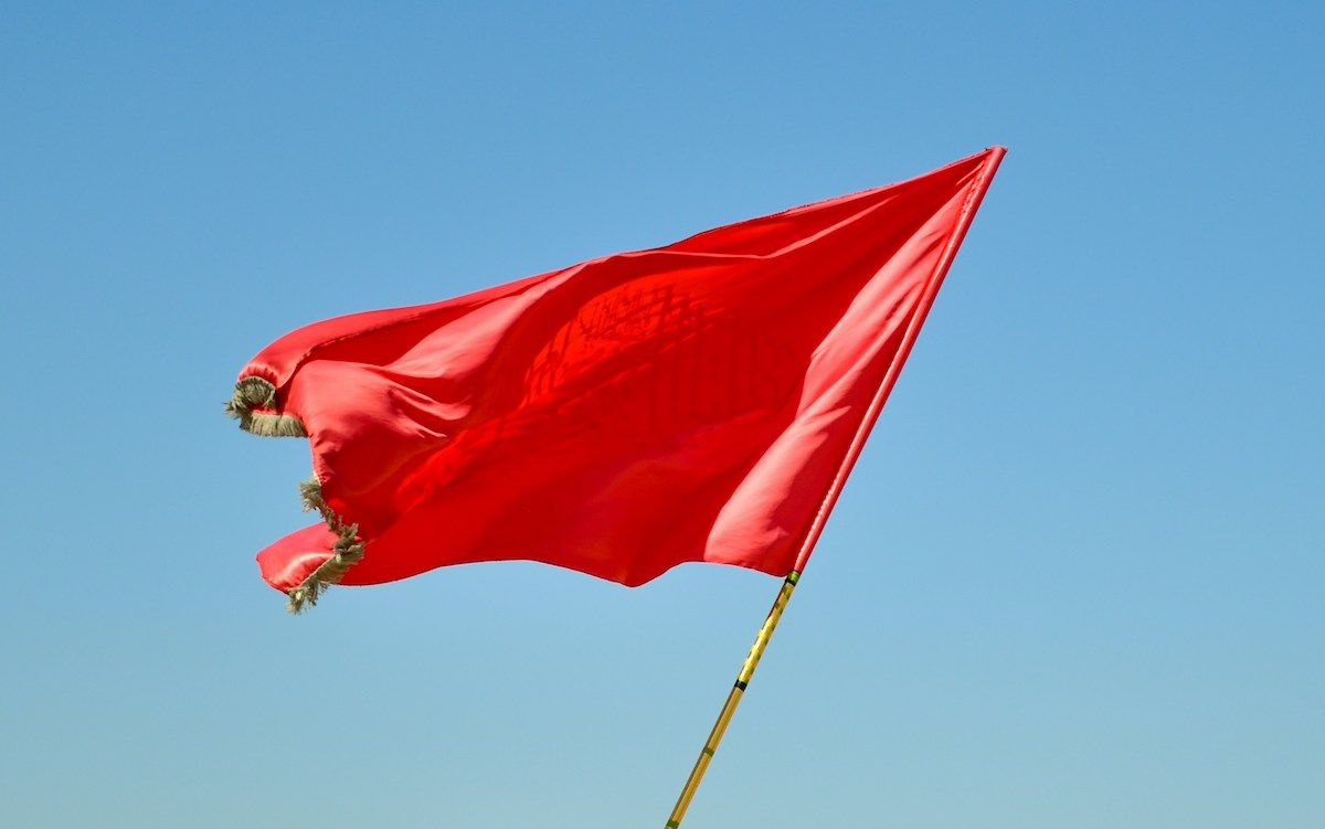 A bright red flag with frayed edges flutters against a clear blue sky. The flag appears slightly tattered at the corners and features a barely visible emblem or design in the center due to lighting and fabric folds.