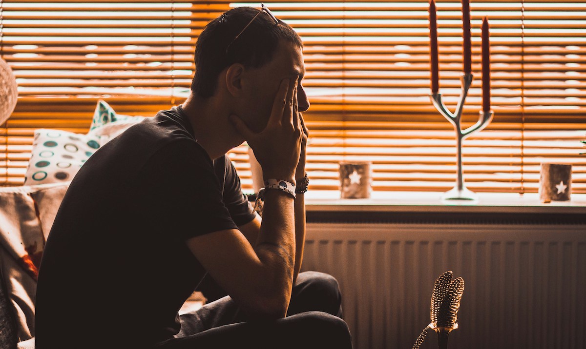 A man sits indoors with his hands covering his face, lit by warm window light filtering through blinds. The scene captures a moment of emotional distress or contemplation.