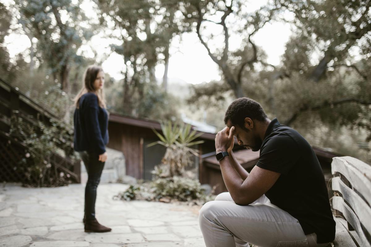 A man sits on a bench outside, hunched forward with his head in his hand, while a woman stands at a distance looking back. The tension in their body language suggests a strained or emotional moment in the relationship.
