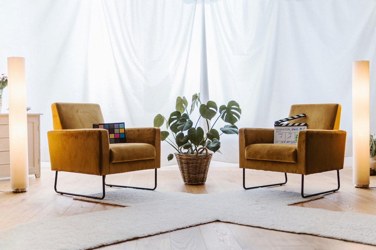Two mustard-yellow armchairs face each other in a bright, airy room, surrounded by houseplants and warm lighting. This cozy setup reflects a classic couples therapy environment, prompting comparisons to digital tools like ChatGPT that some wonder could replace this face-to-face experience.